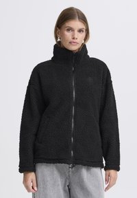 Mustal fleece jakk kõrge kraega ja lukuga. Omab tekstuurset, karvane pinda ja pikki varrukaid.