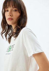 Hvid bomulds T-shirt med korte ærmer, der fremhæver "HOPE" i sort tekst og grøn grafisk design, som fremhæver held-symboler. Glat tekstur.