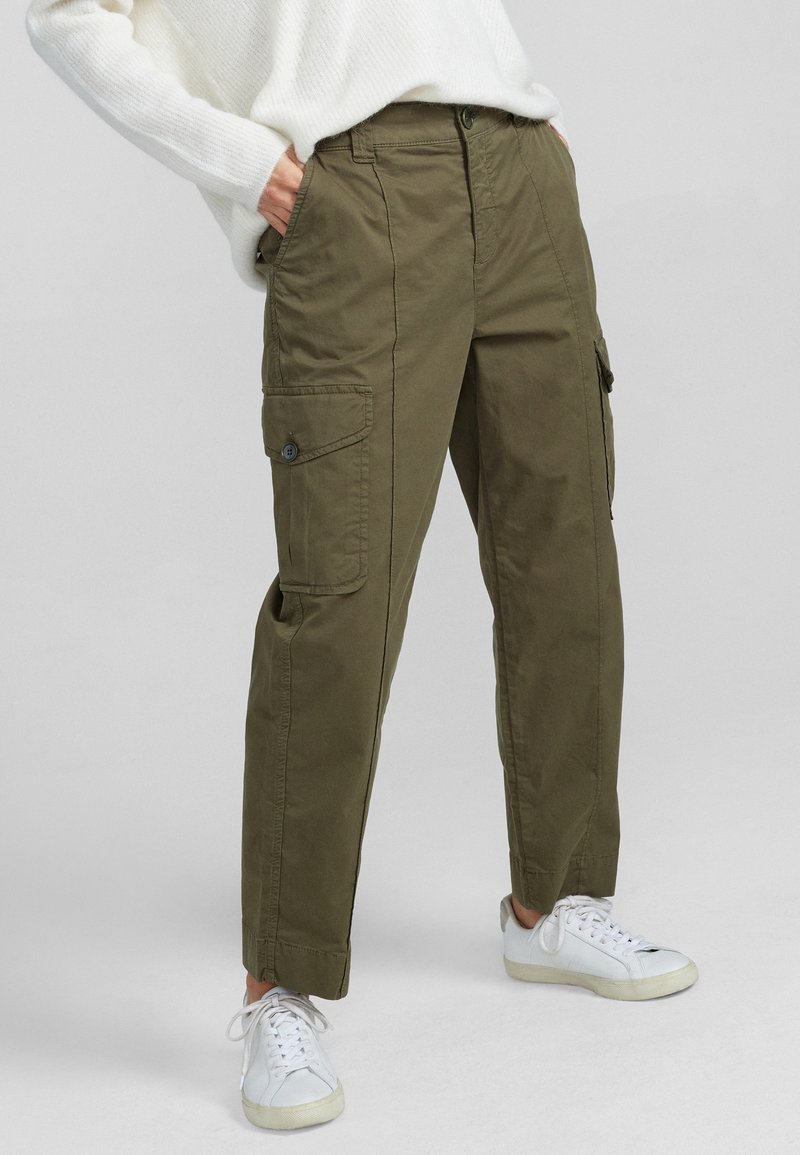 Personne portant un pantalon cargo vert olive avec des poches latérales, un pull en maille blanc et des baskets blanches, debout devant un fond uni.