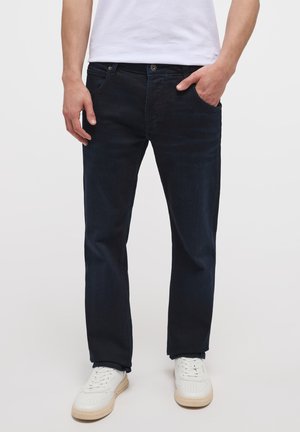 Jeans de mezclilla azul oscuro con corte recto, cinco bolsillos, cierre de botón de latón y una textura sutil, combinados con zapatillas blancas.