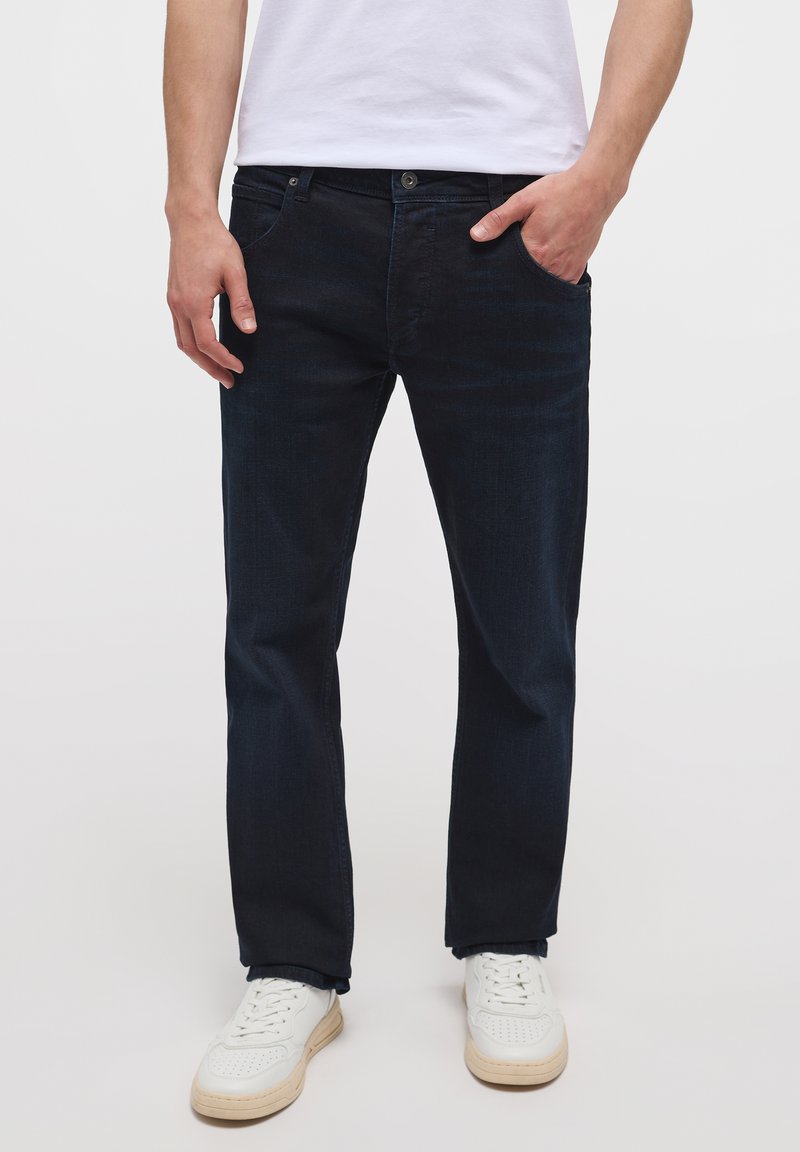 Jeans in denim blu scuro con taglio dritto, cinque tasche, chiusura con bottone in ottone e una texture sottile, abbinati a sneakers bianche.