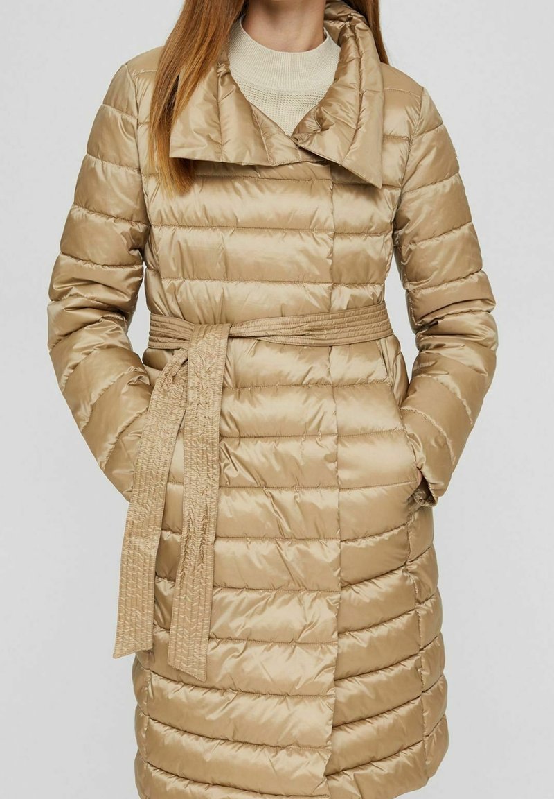 Gepolsterter beiger Mantel mit einem taillierten Gürtel, breiten Revers, horizontaler Steppung und langen Ärmeln. Das Material hat eine glänzende, glatte Textur.