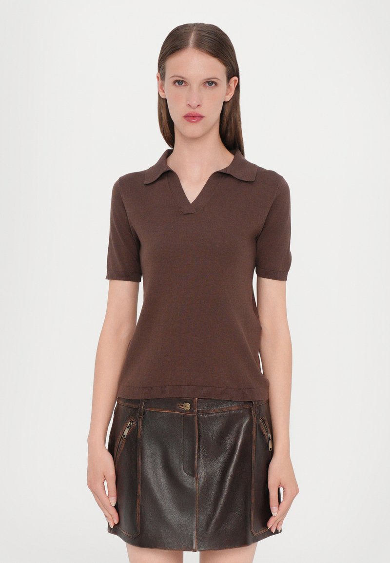 Polo shirt marron à manches courtes avec un col en V, fabriqué en matière douce, présenté au-dessus d'une jupe en cuir marron foncé avec des poches zippées.