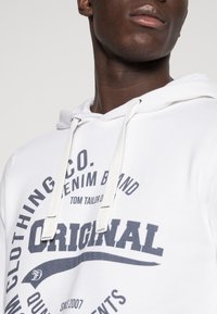 Vit hoodie med marinblå grafik och text, med dragsko i huvan. Materialet verkar mjukt; designen inkluderar varumärkeslogotyp och detaljer.