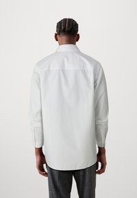 Chemise blanche à boutons, à manches longues, avec couture de pâté de poche au dos et ourlet arrondi. Le tissu est lisse et léger, avec des poignets à un bouton.