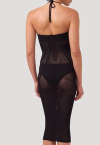 Donna che indossa un abito aderente nero senza spalline in rete con un laccio al collo, con la biancheria intima nera visibile sotto, vista di spalle su uno sfondo semplice.