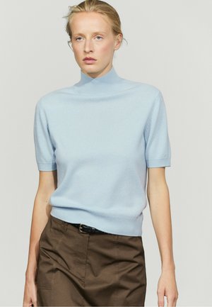 Donna bionda con maglione a dolcevita a maniche corte azzurro chiaro e pantaloni marroni a vita alta su sfondo chiaro uniforme.