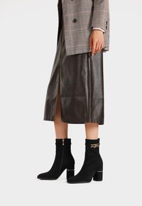 Stivaletti neri in suede con un accento oro, tacco a blocco e cerniera laterale, abbinati a una gonna in pelle marrone e un blazer scozzese.