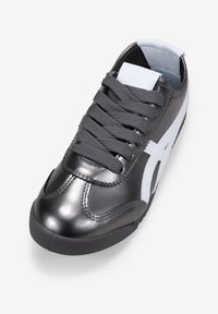 Zapatilla de cuero gris con detalles en blanco, puntera redonda, superficie texturizada y cordones grises. Presenta una lengüeta acolchada y una suela de goma flexible.