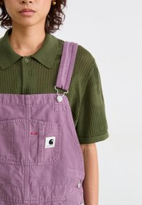 Salopette di denim viola con bretelle regolabili, dotata di una tasca frontale e una toppa con logo. Indossata su una camicia collare verde testurizzata.
