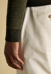 Tume nahk käsi puhkamas kreemikate pükste tasku kõrval, seljas tume roheline tekstuuriga sviiter, mille varrukal on soonikkoes mansetid.