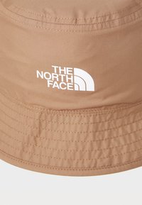 Brązowy kapelusz typu bucket wykonany z lekkiego materiału, z szerokim rondem i białym logo "The North Face" z przodu.
