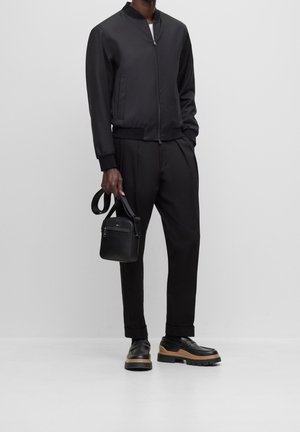 Czarna kurtka typu bomber na zamek, dopasowane czarne spodnie oraz masywne czarne buty z brązową podeszwą; trzyma małą czarną torebkę.