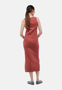 Robe sans manches, longue jusqu'aux chevilles, en tissu côtelé terracotta avec une silhouette ajustée, complétée par des chaussures plates bleues. Vue arrière.