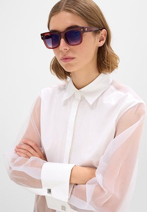 Donna con occhiali da sole sfumati viola e camicia bianca abbottonata con maniche trasparenti, braccia incrociate su sfondo semplice.