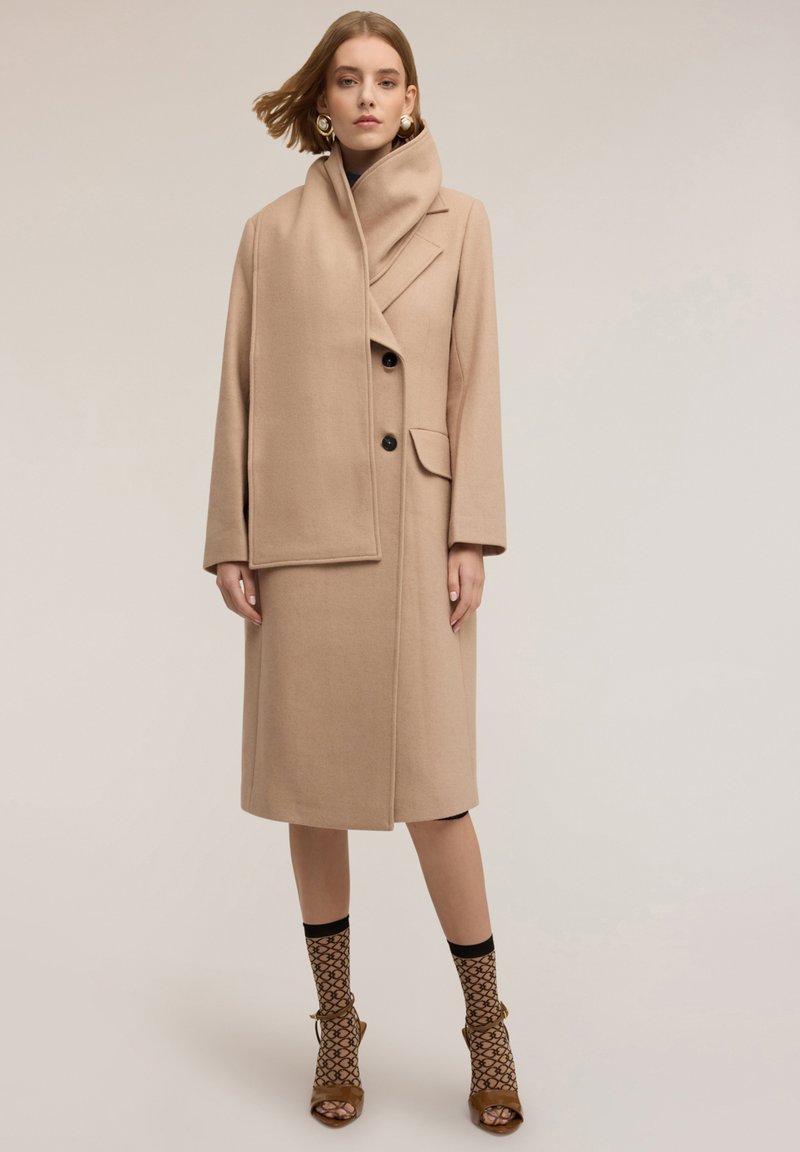 Cappotto di lana beige con colletto alto, design monopetto, bottoni neri e tasche laterali. Il modello indossa collant con fantasia e scarpe con tacco.