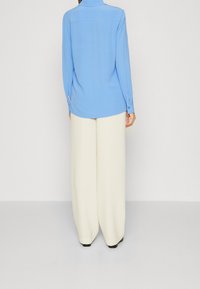 Camicetta azzurro chiaro a maniche lunghe con colletto, polsini con bottone e orlo dritto, abbinata a pantaloni a gamba larga beige.