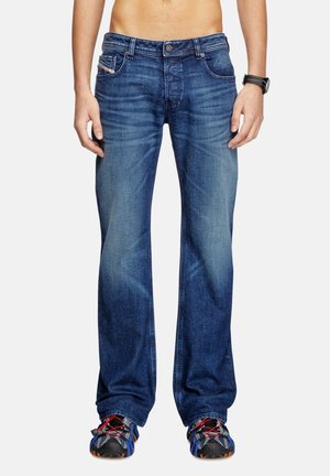 Mann trägt dunkelblaue Jeans mit geradem Bein und ausgewaschenen Details sowie schwarze Sportschuhe mit roten Schnürsenkeln, steht vor einfarbigem Hintergrund.