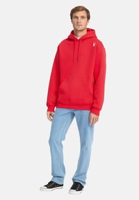Haut à capuche rouge avec une poche avant, capuche à cordon de serrage et logo brodé sur la poitrine, associé à un jean light blue et des baskets noires.