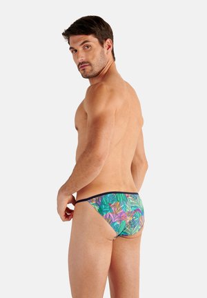 Man met kort donker haar die kleurrijke zwemslips met tropische print draagt, staand met rug en hoofd naar de camera gedraaid.