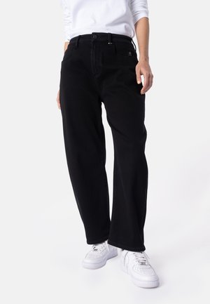 Person iført sorte vidde bukser, hvid langærmet top og hvide sneakers, vist fra taljen og ned mod fødderne mod en ensfarvet baggrund.