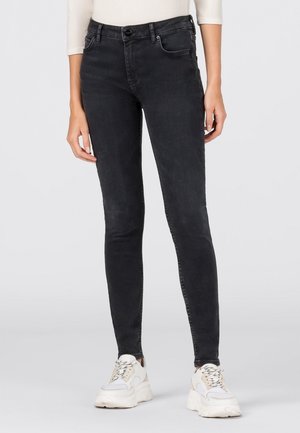 Černé džíny skinny nošené s bílými masivními teniskami a krémovým dlouhým rukávem na jednobarevném pozadí.