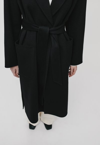 Black wool coat with a belt, featuring two side pockets, a shawl collar, and a structured silhouette. White undershirt and light-colored pants visible.