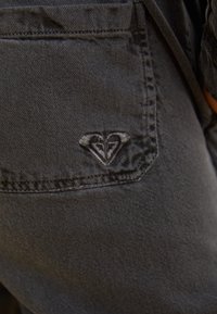 Pantaloni in denim nero con un design a cuore cucito sulla tasca posteriore. Il tessuto sembra resistente e ha una consistenza liscia.