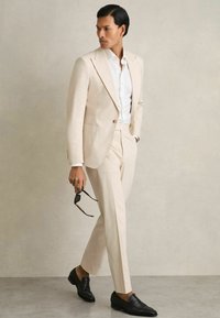Costume beige ajusté avec des revers crantés, associé à une chemise blanche. Mocassins noirs, tenant des lunettes de soleil, contre un fond neutre.
