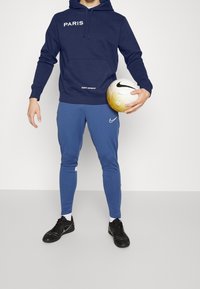 Granatowy hoodie z napisem "PARIS" i "SAINT-GERMAIN", w zestawieniu z niebieskimi dopasowanymi spodniami dresowymi i czarnymi butami sportowymi. Model trzyma piłkę nożną z złotymi akcentami.