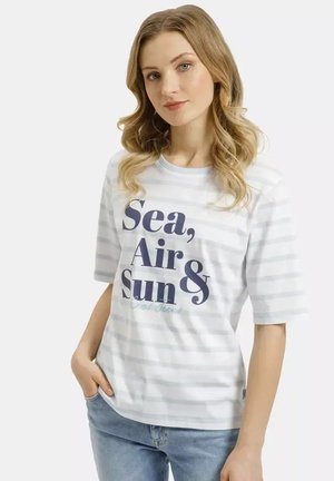 Młoda kobieta o blond, falowanych włosach, ubrana w biały, w paski t-shirt z napisem „Sea, Air & Sun”, pozująca z jedną ręką w kieszeni niebieskich dżinsów.