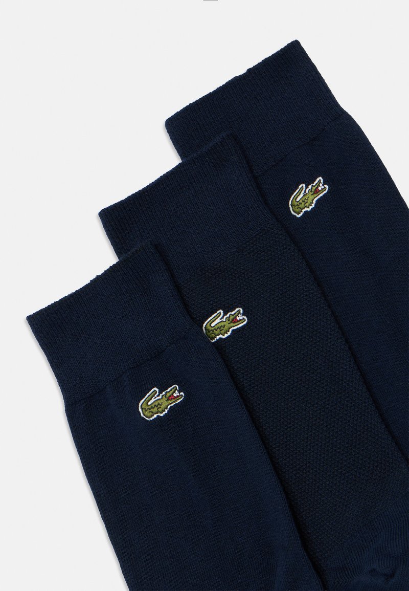 Calcetines azul marino con patrones ribeteados y texturizados, con un pequeño logo de cocodrilo verde bordado cerca del puño. Se muestran tres pares.