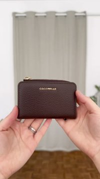 Brūns ādas maciņš ar teksturētu apdari, zelta "COCCINELLE" logo, ar rāvējslēdzēju un kompakta, taisnstūra forma.