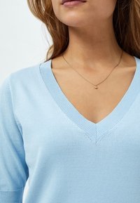 Světle modrý pletený svetr s výstřihem do V a krátkými rukávy, s vroubkovanými okraji. Na krku je viditelný jemný zlatý náhrdelník s malým přívěskem.