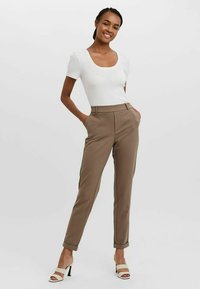 Bruine op maat gemaakte broek met een hoge taille, omslagnaden aan de broekspijpen en een geweven witte top met korte mouwen. Gepaarde met witte haksandalen.