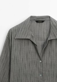 Camicia grigia con colletto, decorata con righe verticali di diverse tonalità, caratterizzata da una superficie liscia e bottoni azzurri per la chiusura.