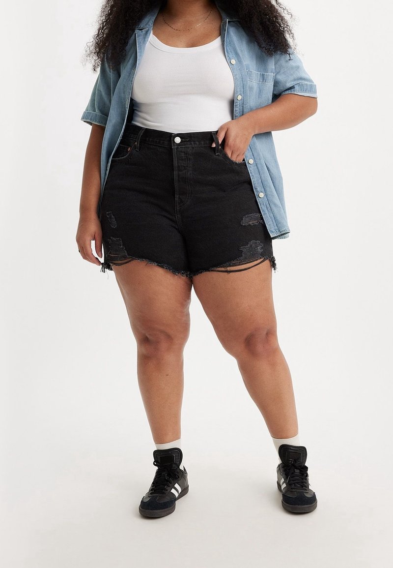 Femme portant un short en jean noir déchiré taille haute, un débardeur blanc, une chemise à manches courtes bleu clair boutonnée et des baskets noires.