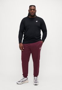 Camiseta negra de manga larga con logo, combinada con pantalones de chándal burdeos con puños elásticos. Los zapatos son de color gris claro con acentos más oscuros.