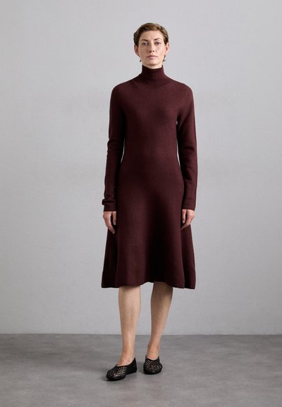 Abito borgogna a maniche lunghe con collo alto, gonna a campana, realizzato in morbido tessuto a maglia. Indossato con scarpe slip-on nere perforate.