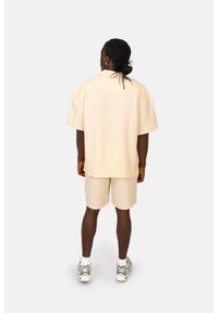 Camicia leggera beige a maniche corte con vestibilità comoda, abbinata a pantaloni corti coordinati. Tessuto morbido con una sottile trama; indossata con sneakers grigie.