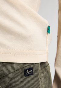 Beige katoenen shirt over olijfgroene, gestructureerde broek. Opvallend label: "Petrol Industries" gestikt op de broek.