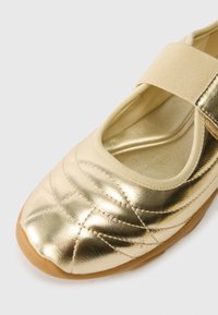 Zeltītas ādas Mary Jane kurpes ar quilted rakstu, elastīgu jostu un gaiši brūnu gumijas zoli. Gluda tekstūra ar spīguļojošu apdari.