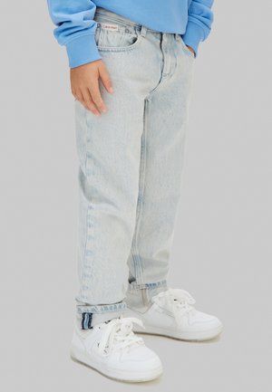 Enfant portant un jean bleu clair, un sweat-shirt bleu et des baskets blanches, debout avec une main détendue le long du corps et l'autre dans la poche.