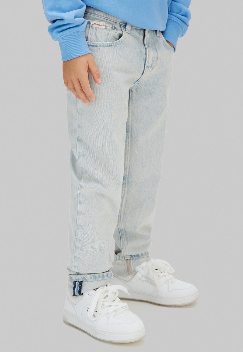 Παιδί που φοράει ανοιχτό μπλε τζιν, μπλε φούτερ και λευκά αθλητικά παπούτσια, στέκεται με το ένα χέρι χαλαρό στο πλευρό και το άλλο στην τσέπη.