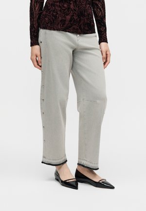 Woman wearing light gray, wide-leg pants with snap buttons along the sides, black pointed flats, and a long-sleeve patterned top.