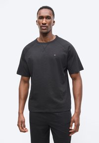 Άνδρας που φοράει απλό μαύρο κοντομάνικο πουκάμισο και μαύρο παντελόνι, στέκεται με τα χέρια χαλαρά σε λευκό απλό φόντο.