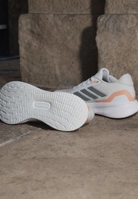 Baskets athlétiques blanches et pêche avec des rayures grises, l'une montrant le profil latéral et l'autre la semelle vers le haut sur une surface en pierre.