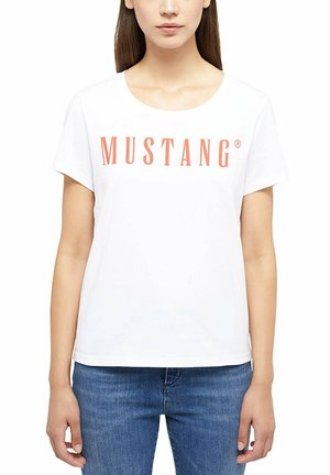 Frau, die ein weißes T-Shirt mit kurzen Ärmeln mit orangefarbenem "MUSTANG"-Schriftzug und blaue Jeans trägt, steht vor einem schlichten weißen Hintergrund.