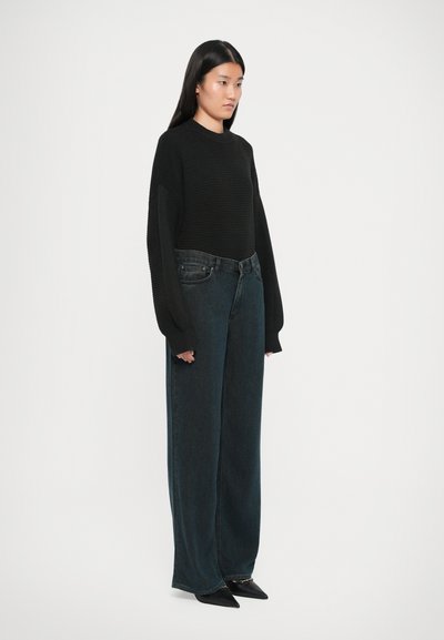 Suéter negro de punto con mangas largas, combinado con unos jeans oscuros de corte holgado y zapatos negros de punta, mostrado en vista lateral.