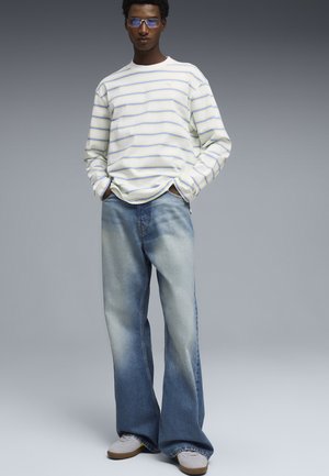 Mann mit blau getönten Brillen, weißem Langarmhemd mit hellblauen Streifen, weit geschnittenen, ausgewaschenen Jeans und weißen Turnschuhen, stehend mit den Händen in den Taschen.